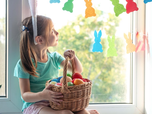 Une petite fille avec des oreilles de lapin sur le rebord de la fenêtre sourit dans les mains d'un panier avec des oeufs de Pâques