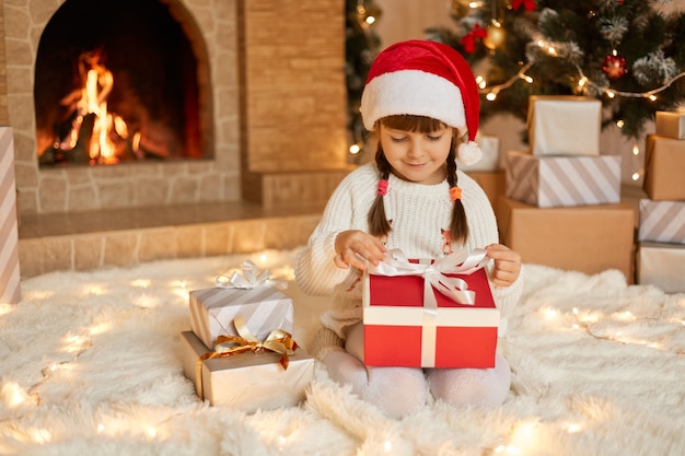 Petite fille ouvrant le cadeau de Noël, gardant les doigts sur les rubans, regardant la boîte-cadeau avec le sourire, enfant portant un chapeau de père Noël posant sur le sol près de la cheminée et de l'arbre de Noël.