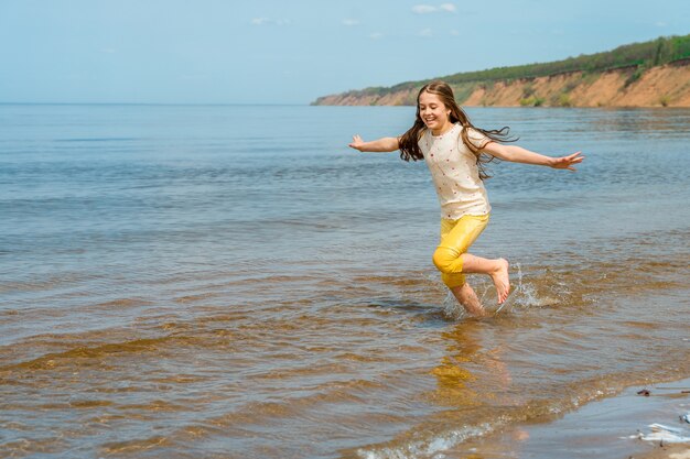 Une petite fille en pantalon jaune vif court dans l'eau et s'ébat sur la plage