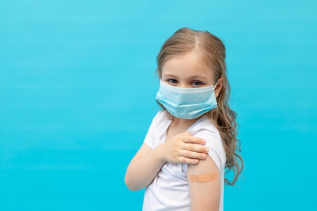 Une petite fille avec un patch sur son bras après la vaccination dans un masque de protection médicale dans un t-shirt blanc sur fond bleu dans l'espace de studio pour le texte le concept de santé vaccinale