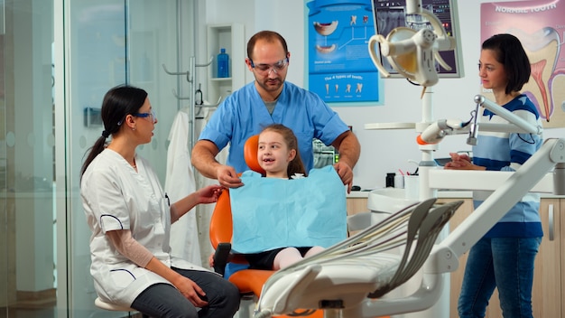 Petite Fille Patiente Souffrant De Douleurs Dentaires Expliquant Le Problème Dentaire Au Dentiste Pédiatrique Et Indiquant Un Mal De Dents Avec La Langue. Stomatologue Prenant Avec La Mère Au Sujet De L'examen Stomatologique De L'enfant.