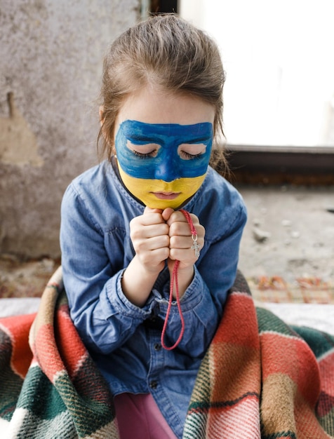 Une petite fille patriote ukrainienne avec le drapeau de l'Ukraine sur son visage prie assise à la fenêtre