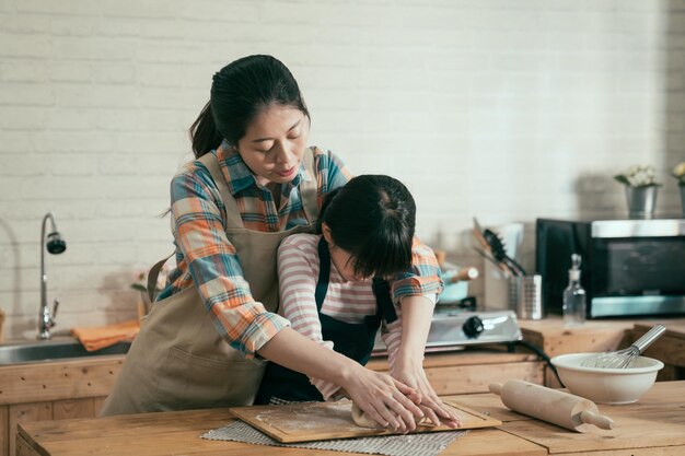 Petite fille pétrissant de la farine avec une maman asiatique le jour des mères gâteau fait main pour pâques. jeune femme en tablier enseignant à un enfant la cuisson du pain pour l'anniversaire de papa. la famille aime passer du temps ensemble dans la cuisine de la maison