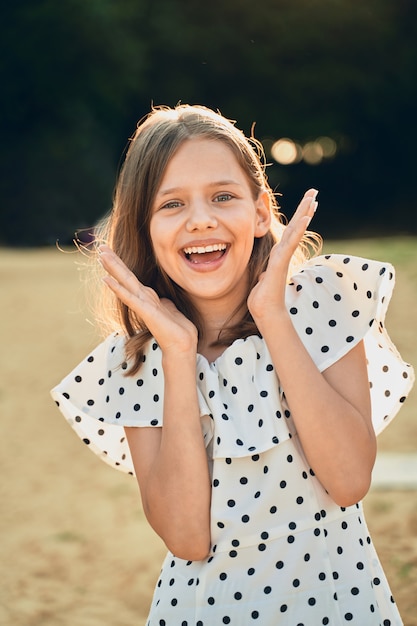 Une petite fille sur la plage dans une robe à pois rit et met ses mains sur le côté.