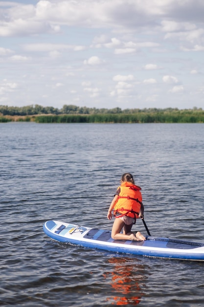 Petite fille sur une planche sup agenouillée regardant la surface de l'eau avec une flaque d'eau dans la main droite mise dans l'eau portant un gilet de sauvetage Vacances actives pleines d'aventures Inculcation de l'amour pour le sport depuis l'enfance