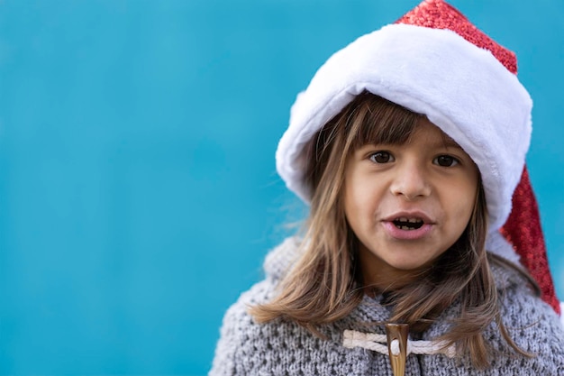 Petite fille portant un chapeau de père Noël regardant la caméra Drôle enfant jouant contre un fond de mur bleu à l'extérieur au moment de Noël Joyeux Noël et bonne année