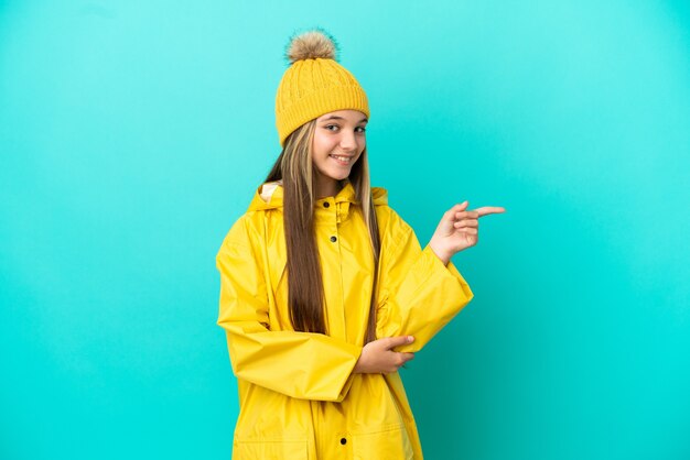 Petite fille portant un manteau imperméable sur fond bleu isolé, pointant le doigt sur le côté