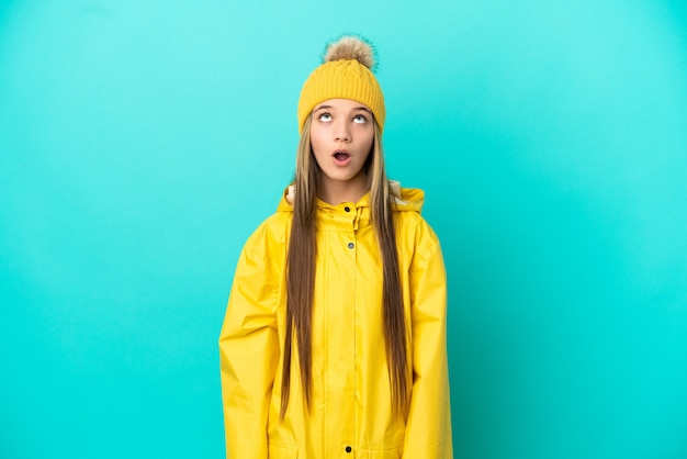 Petite fille portant un manteau imperméable sur une surface bleue isolée en levant et avec une expression surprise