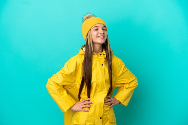 Petite fille portant un manteau imperméable sur une surface bleue isolée posant avec les bras à la hanche et souriant