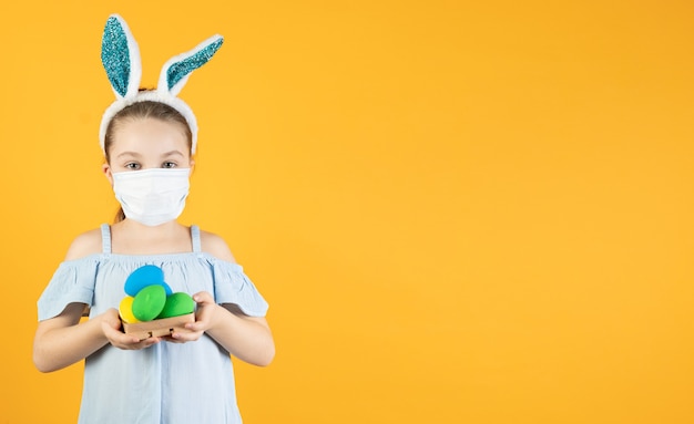 Une petite fille portant un masque médical de coronavirus sur son visage, sur sa tête avec des oreilles de lapin, tient des œufs de Pâques de différentes couleurs dans ses mains
