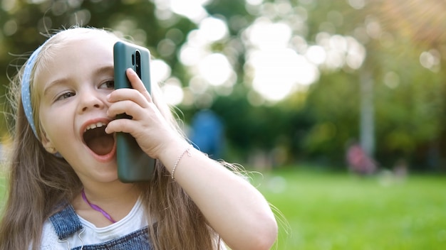 Petite fille positive ayant une conversation sur son téléphone portable dans un parc d'été. Jeune fille parlant au sellphone.