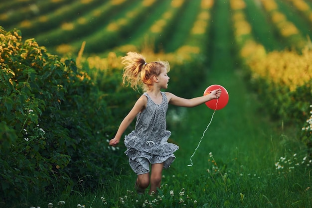 Une petite fille positive avec un ballon rouge dans les mains s'amuse sur le terrain pendant la journée d'été
