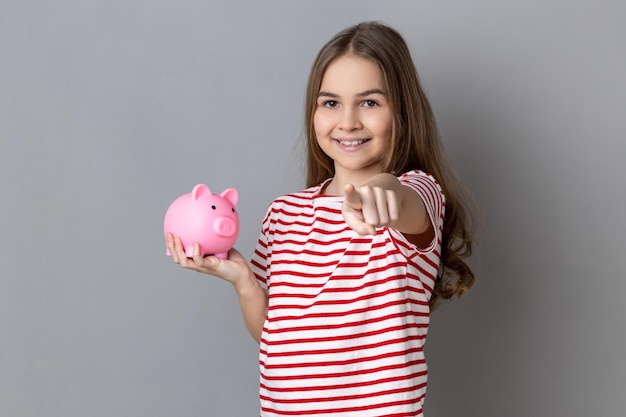 Petite fille positive debout pointant vers la caméra tenant une tirelire économiser de l'argent bancaire