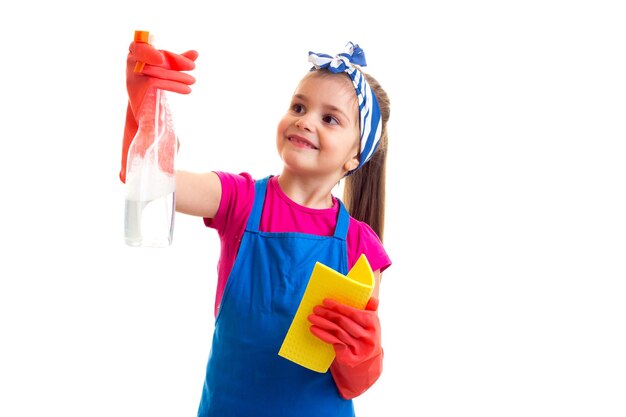 Petite fille positive avec queue de cheval en tablier bleu avec des gants rouges tenant un spray et un plumeau jaune