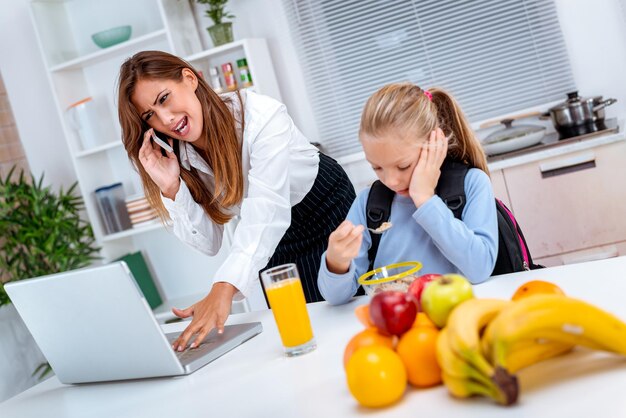 Une petite fille prenant un petit-déjeuner pendant que sa mère surmenée téléphonait et vérifiait quelque chose sur son ordinateur portable avant d'aller au bureau. La mère se met en colère à cause d'un stress.