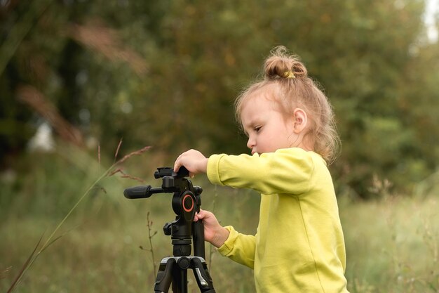 petite fille préscolaire curieuse aux cheveux blonds ondulés jouant avec un trépied photo dans le parc