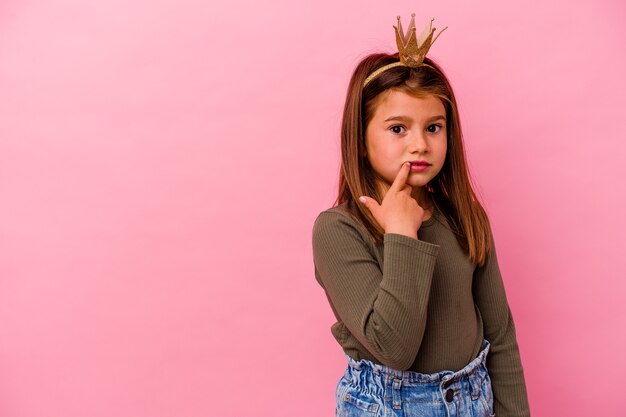 Petite fille princesse avec couronne isolée sur rose pensée détendue sur quelque chose en regardant un espace de copie.