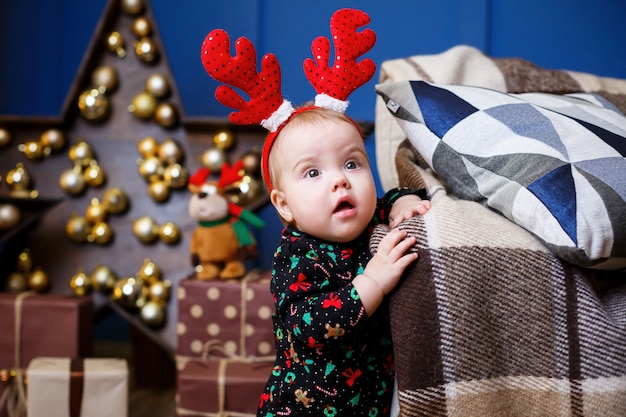 Une petite fille en pull chaud est assise sous un arbre de Noël avec des jouets et des cadeaux avec des cornes sur la tête. Enfance heureuse. Ambiance de vacances du nouvel an