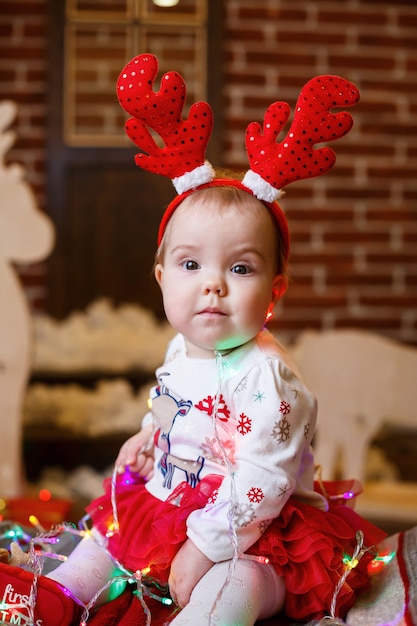 Une petite fille en pull chaud est assise sous un arbre de Noël avec des jouets et des cadeaux avec des cornes sur la tête. Enfance heureuse. Ambiance de vacances du nouvel an