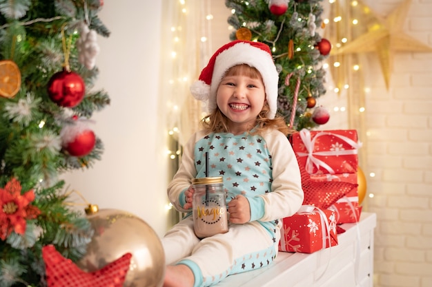 Petite fille en pyjama et bonnet de Noel buvant du lait de cacao