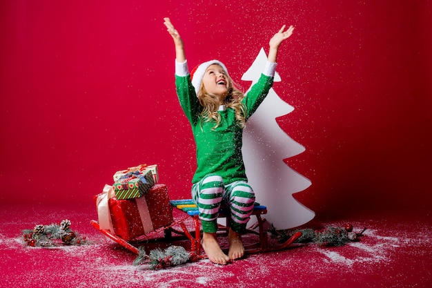 petite fille en pyjama de Noël ou costume d'elfe et bonnet de noel attrape la neige assis sur un traîneau