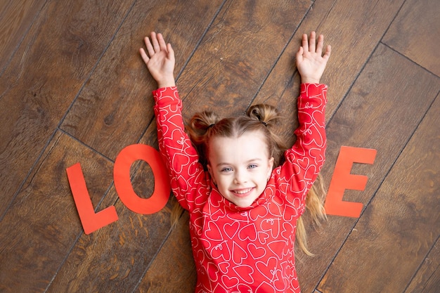 une petite fille en pyjama rouge avec l'inscription Love se trouve sur un fond de bois brun foncé sur le sol et rit en tenant ses mains en forme de la lettre V Jour de la Saint-Valentin