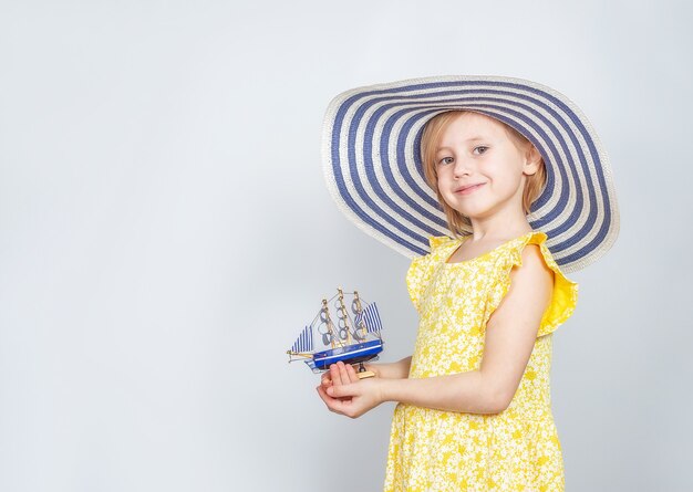 Une petite fille de race blanche dans un chapeau à larges bords tient un bateau jouet dans ses mains. Le concept des vacances d'été