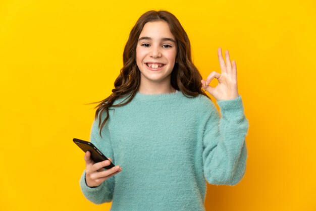 Petite fille de race blanche isolée sur un mur jaune à l'aide de téléphone mobile et faisant signe OK