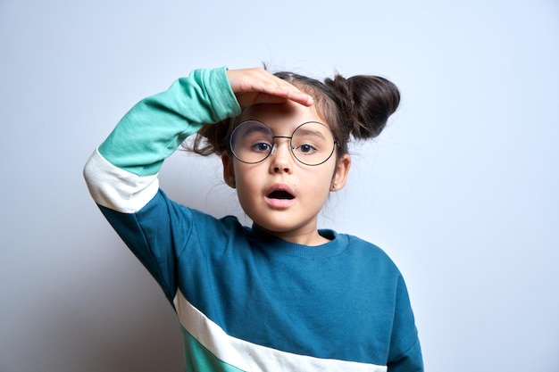 Petite fille de race blanche avec des lunettes regarde l'avenir à la distance de loin avec la main sur la tête isolé sur fond blanc Concept de recherche