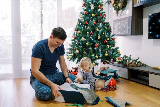 La petite fille regarde avec la bouche ouverte son père déballer les cadeaux près de l'arbre de Noël.