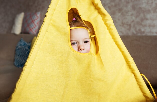 Une petite fille regarde par la fenêtre d'un tipi, joue à la maison sur le canapé. Jeux d'enfants à la maison.