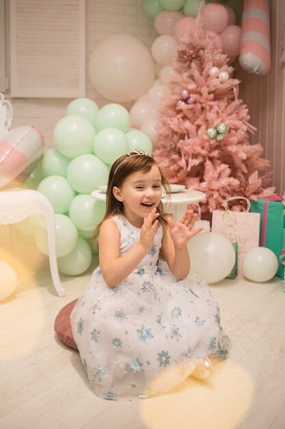 Une petite fille riante vêtue d'une robe moelleuse est assise sur des oreillers et frappe des mains dans les décorations du Nouvel An