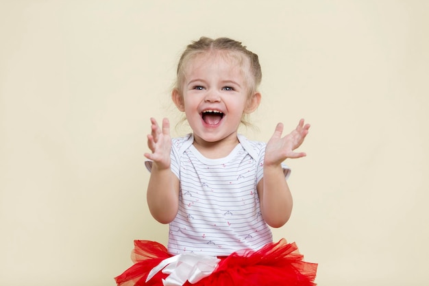 La petite fille rit joyeusement et tape dans ses mains Joli bébé dans une jupe rouge avec une banque et un t-shirt blanc Enfance heureuse et émotions sincères Fond jaune