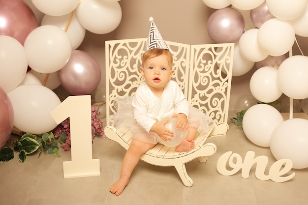 petite fille en robe blanche assise sur un banc blanc anniversaire numéro un avec des ballons