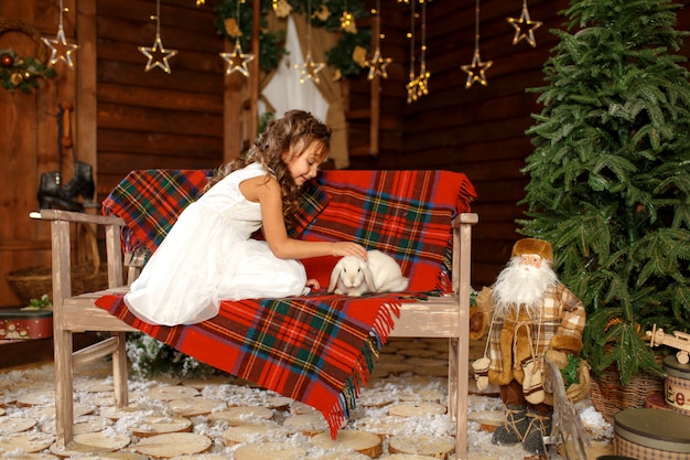 Une petite fille en robe blanche assise sur le banc avec un lapin blanc.