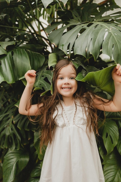 Petite fille en robe blanche dans un jardin botanique un enfant se tient près des feuilles de Monstera