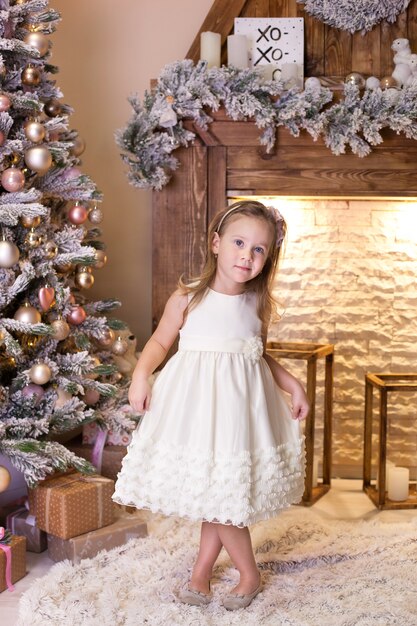 Petite fille en robe blanche se tient près de l'arbre de Noël décoré et cheminée dans le salon