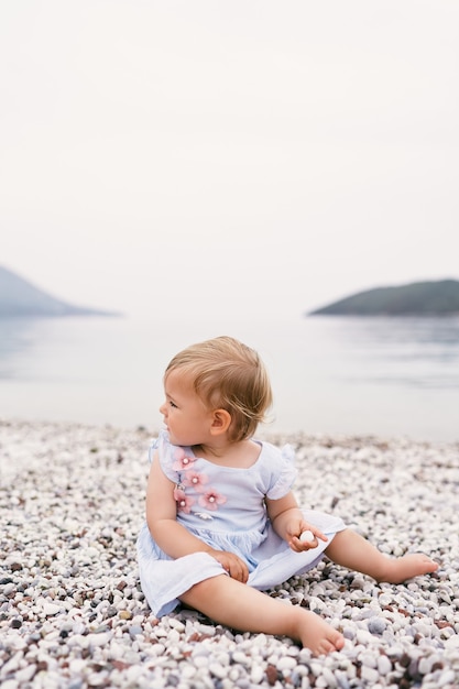 Petite fille en robe est assise sur une plage de galets en tournant la tête en arrière