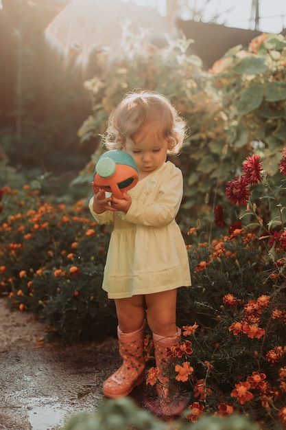 Petite fille en robe jaune et bottes en caoutchouc arrose des fleurs dans le jardin Petite aide