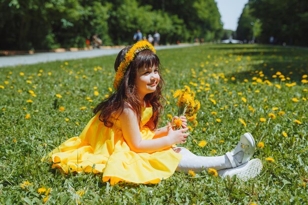 Une petite fille en robe jaune et avec une couronne de pissenlits sur la tête est assise sur l'herbe du parc