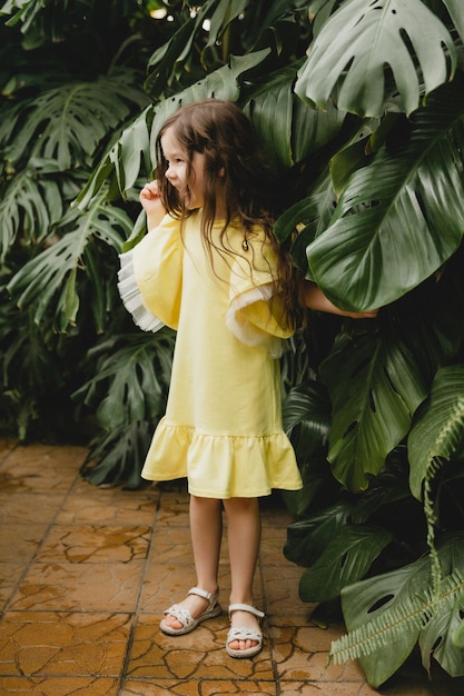Petite fille en robe jaune dans un jardin botanique un enfant se tient près des feuilles de Monstera