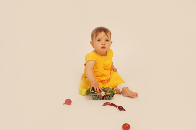 petite fille en robe jaune est assise sur un fond blanc à côté d'un petit panier avec des légumes