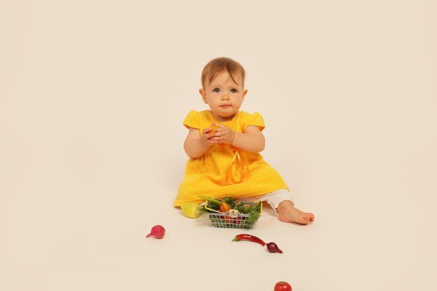 petite fille en robe jaune est assise sur un fond blanc à côté d'un petit panier avec des légumes