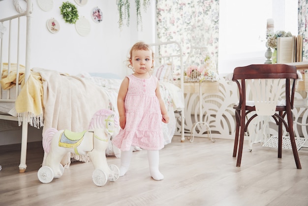 Petite fille en robe mignonne implantée au lit jouant avec des jouets à la maison. Chambre d'enfant vintage blanche. Concept d'enfance