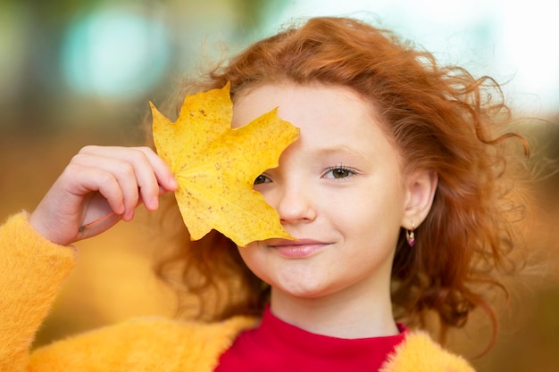 Petite fille rousse ferme un œil avec une feuille d'érable jaune Enfant en automne jour gros plan