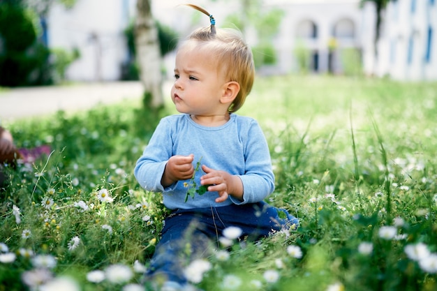 La petite fille s'assied sur l'herbe verte et tient une fleur dans sa main
