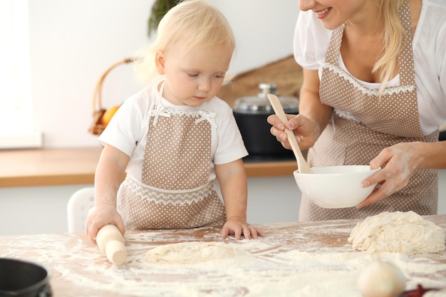 Petite fille et sa maman blonde en tabliers beiges jouant et riant tout en pétrissant la pâte dans la cuisine. Pâtisserie maison pour pain, pizza ou biscuits au four. Concept de plaisir et de cuisine en famille.