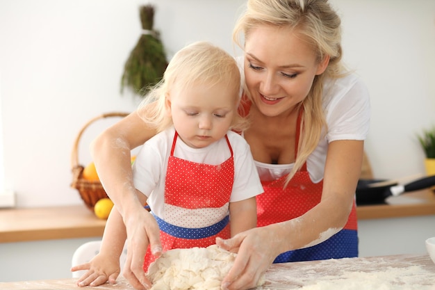 Petite fille et sa maman blonde en tabliers rouges jouant et riant tout en pétrissant la pâte dans la cuisine. Pâtisserie maison pour pain, pizza ou biscuits au four. Concept de plaisir et de cuisine en famille.