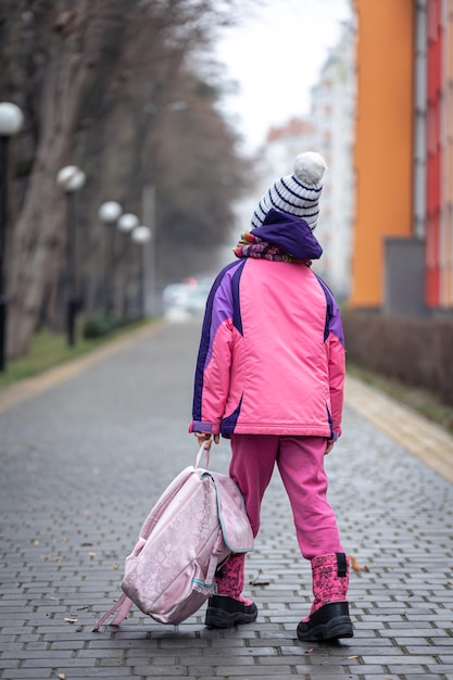 Petite fille avec un sac à dos dans une veste et un chapeau près de l'école