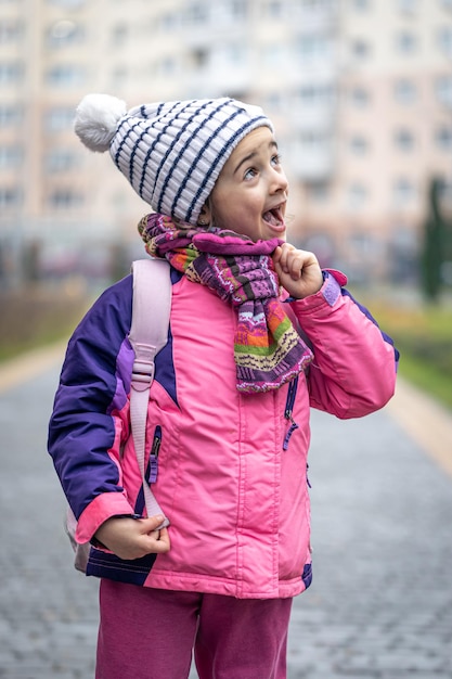 Petite fille avec un sac à dos dans une veste et un chapeau près de l'école